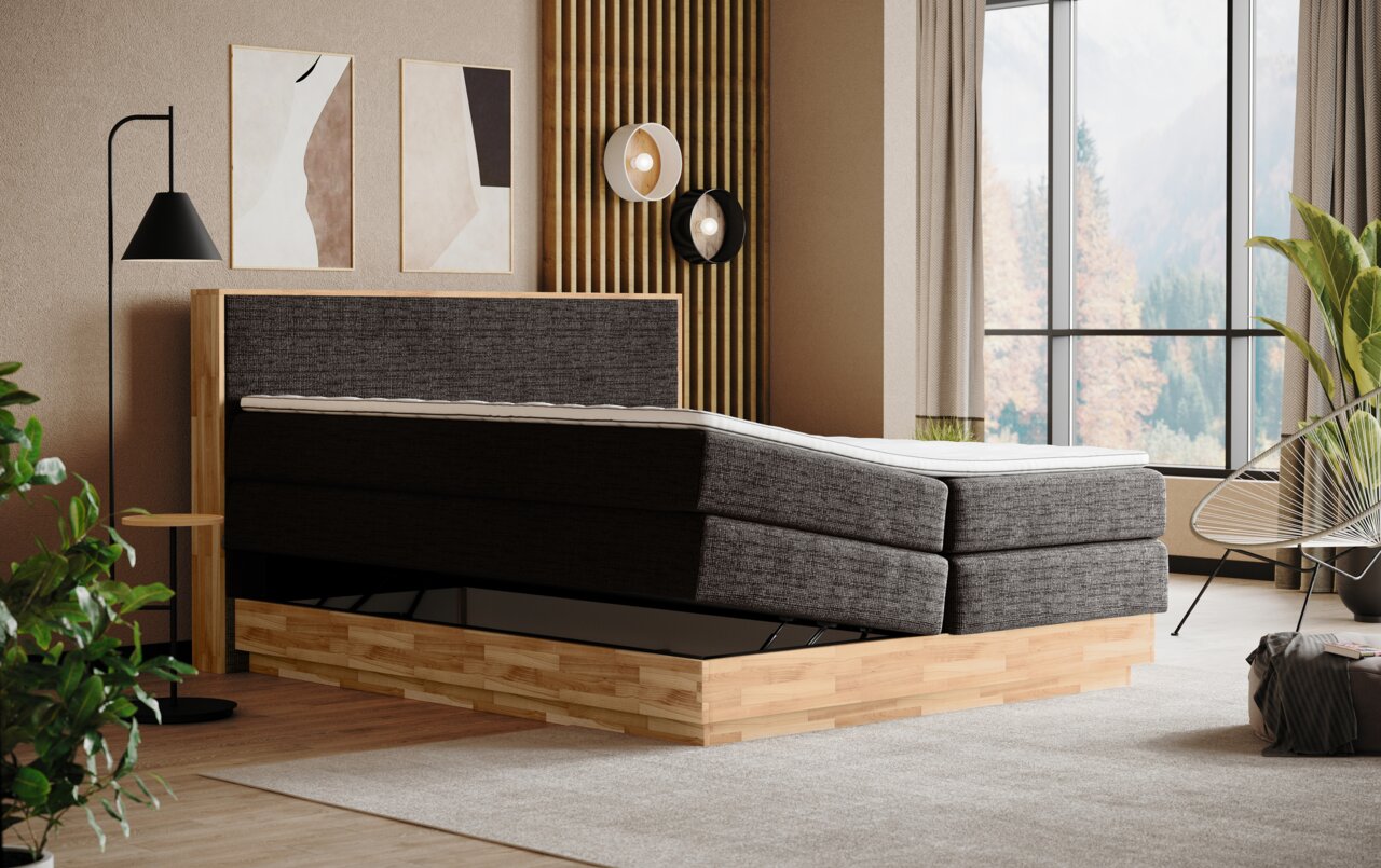 Zijaanzicht van een modern boxspringbed met grijze stoffen bekleding en een houten frame in een stijlvol ingerichte slaapkamer met grote ramen.