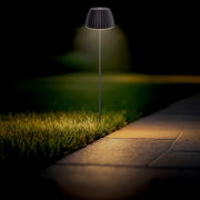 Schwarze LED-Garten-Wegeleuchte mit geriffeltem Schirm auf schlankem Stab, warmes Licht am Rand, am Rasen neben einem Gehweg bei Dämmerung, frontale Perspektive auf Augenhöhe