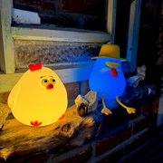 Zwei leuchtende Nachtlichter in Form von Enten, eine gelb mit rotem Kamm und die andere blau mit Hut, auf einem Holzbrett vor einem Fensterbrett platziert. Perspektive von vorne.