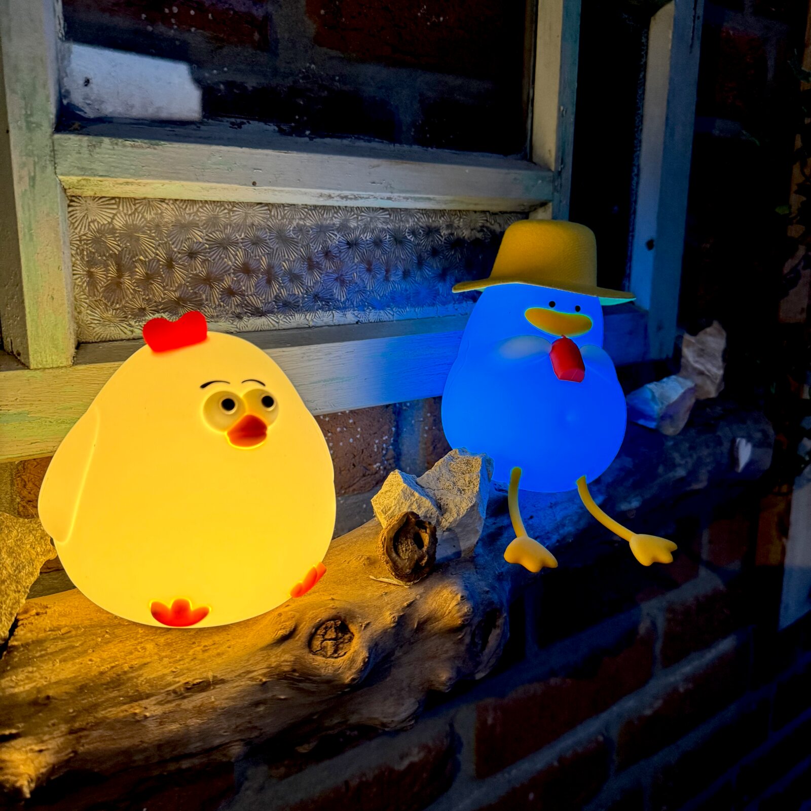 Zwei leuchtende Nachtlichter in Form von Enten, eine gelb mit rotem Kamm und die andere blau mit Hut, auf einem Holzbrett vor einem Fensterbrett platziert. Perspektive von vorne.