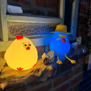 Zwei leuchtende Nachtlichter in Form von Enten, eine gelb mit rotem Kamm und die andere blau mit Hut, auf einem Holzbrett vor einem Fensterbrett platziert. Perspektive von vorne.