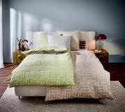 Modernes Bett mit zweifarbiger Bettwäsche in Salbeigrün und Beige mit grafischem Muster und passenden Kissen, frontal aus leicht erhöhter Perspektive im Schlafzimmer-Setting mit Nachttischen und roter Tischlampe