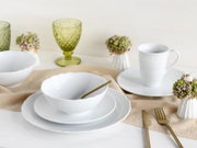 Weißes Porzellan-Geschirrset mit strukturierter Oberfläche (Schale auf Teller, Tasse auf Untertasse) auf hellem Tischläufer, daneben goldenes Besteck und grüne Gläser, fotografiert in leicht erhöhter Frontansicht.