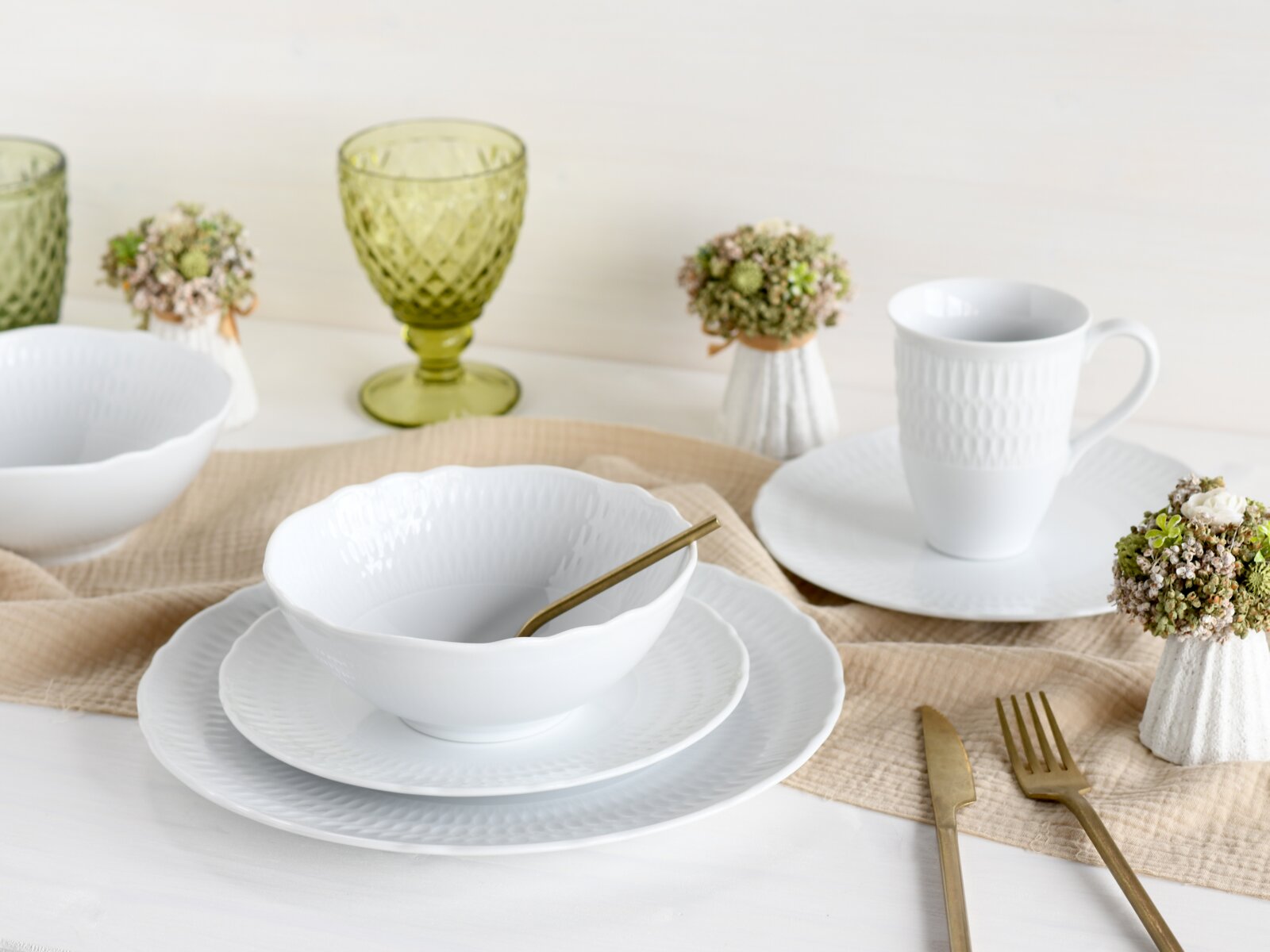 Weißes Porzellan-Geschirrset mit strukturierter Oberfläche (Schale auf Teller, Tasse auf Untertasse) auf hellem Tischläufer, daneben goldenes Besteck und grüne Gläser, fotografiert in leicht erhöhter Frontansicht.
