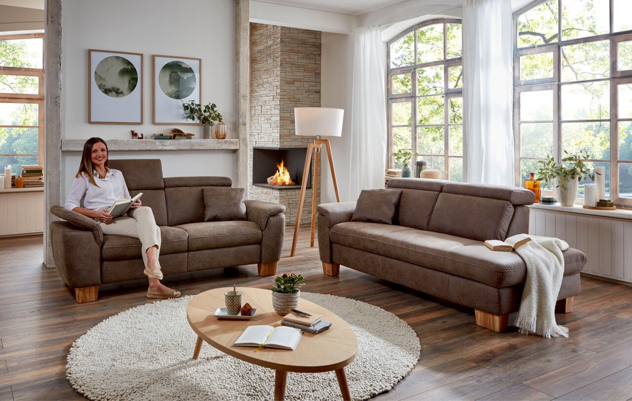 Gemütliches Wohnzimmer mit zwei braunen Sofas, eines davon mit einer Frau, die ein Buch liest. Im Hintergrund ein Kamin und große Fenster mit Blick ins Grüne. Perspektive von vorne.