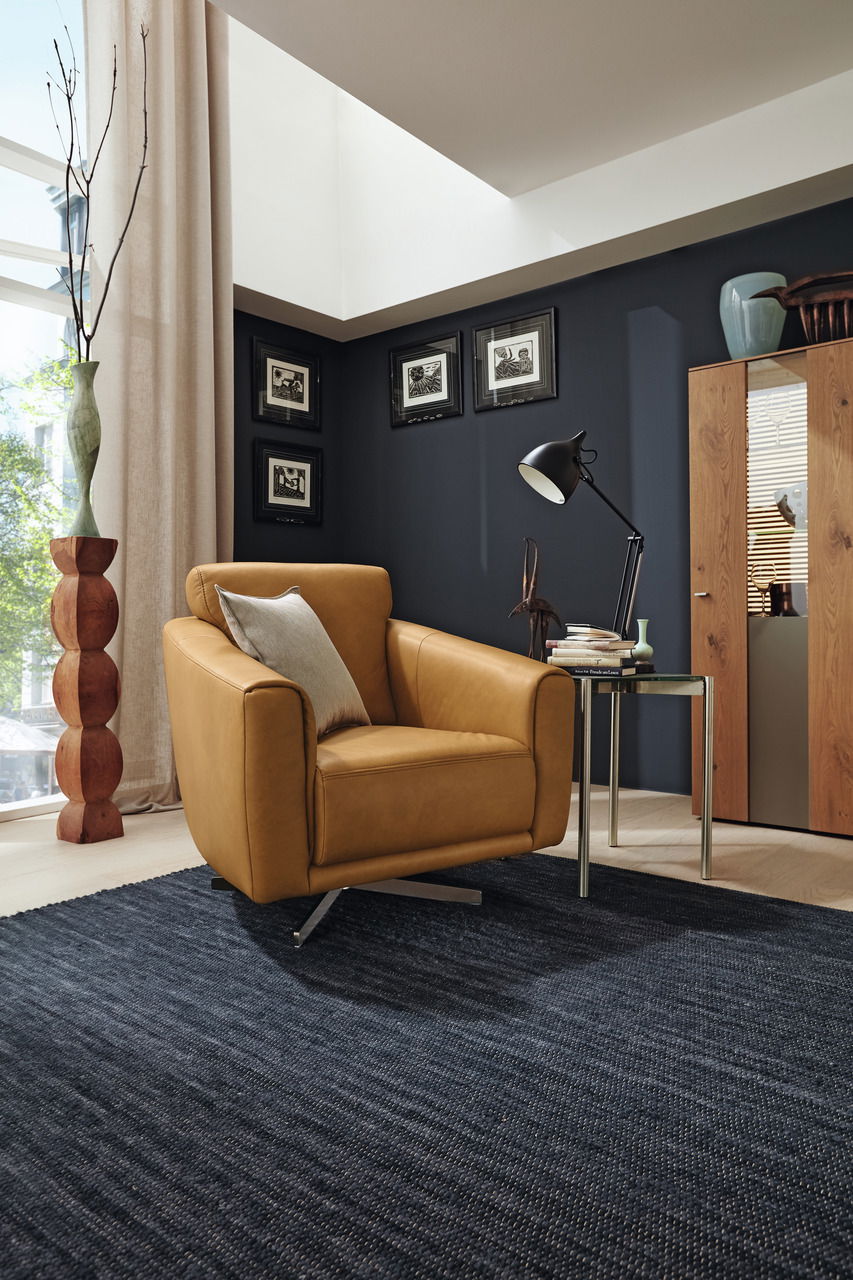 Moderner Drehsessel in senfgelber Farbe, seitliche Perspektive, in einem stilvoll eingerichteten Wohnzimmer mit dunkler Wand und Holzmöbeln.