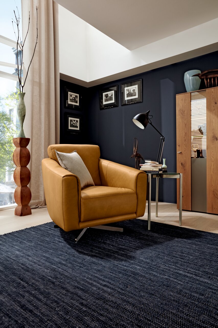 Moderner Drehsessel in senfgelber Farbe, seitliche Perspektive, in einem stilvoll eingerichteten Wohnzimmer mit dunkler Wand und Holzmöbeln.