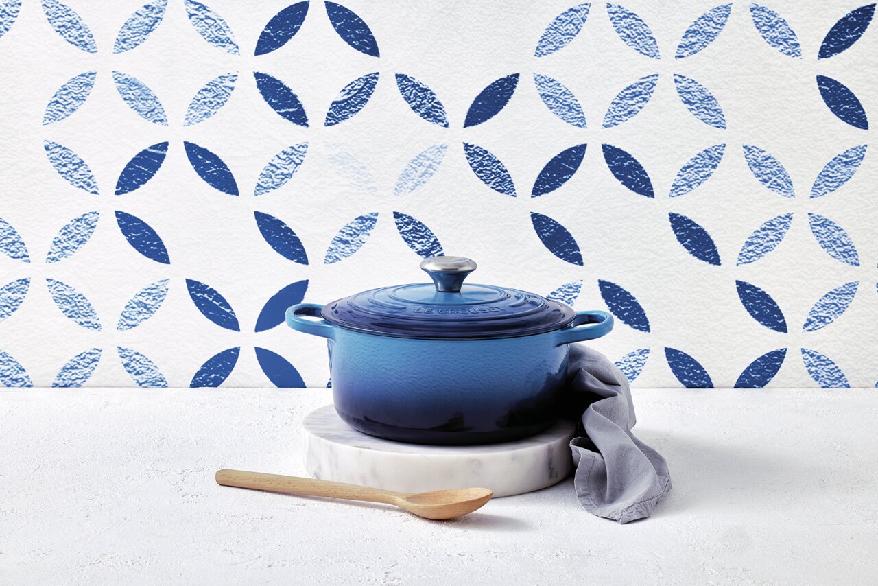 Ronde blauwe braadpan met deksel, geplaatst op een witte marmeren voet, met daarnaast een houten lepel en een grijze doek, gefotografeerd vanuit een zijaanzicht.