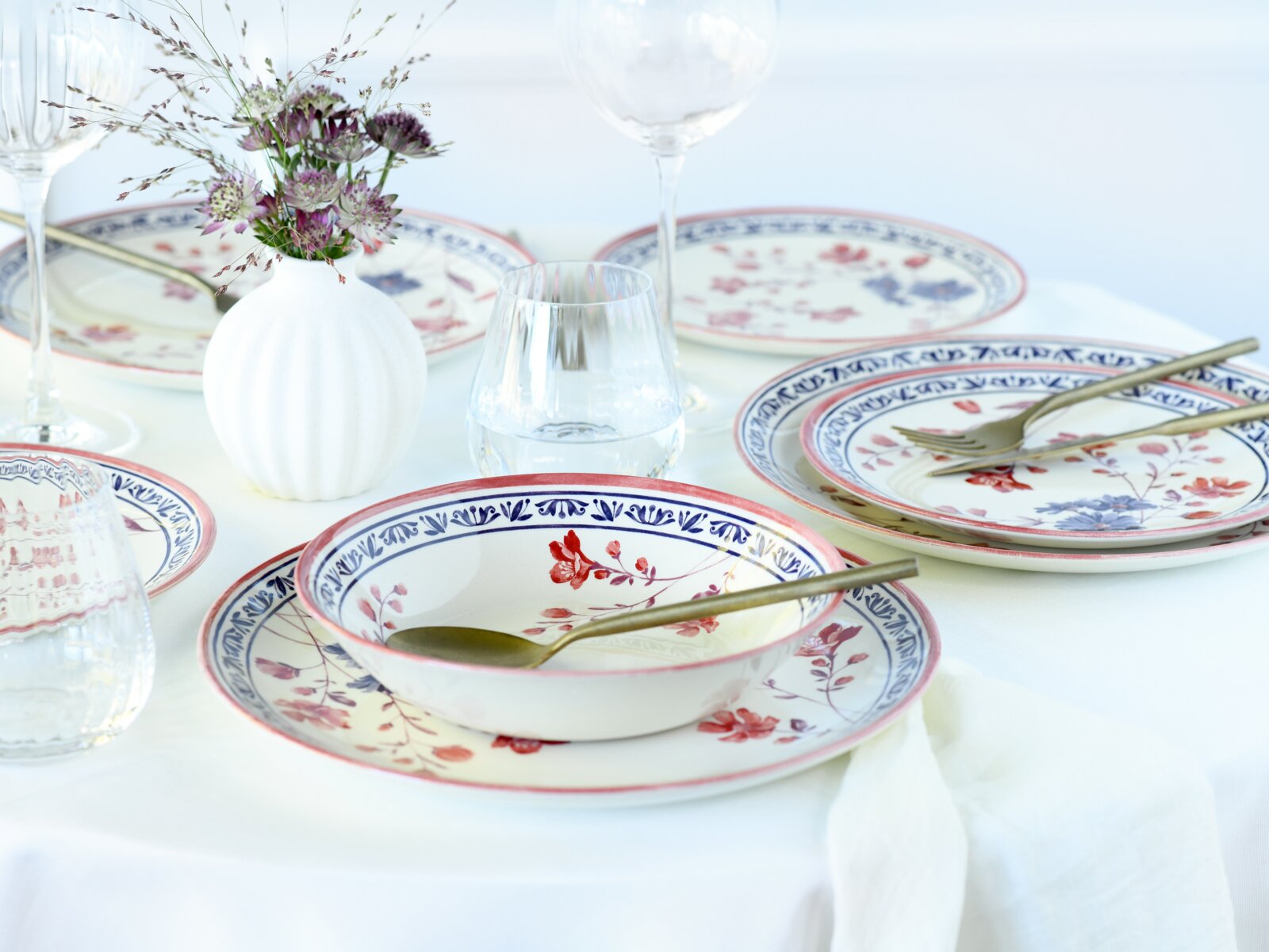 Gedeckter Tisch mit Porzellan-Tellern und Schalen im Blumenmuster in Blau und Rot, goldfarbenem Besteck, Glasvasen und Weingläsern, fotografiert aus leicht erhöhter schräger Perspektive (3/4-Ansicht).