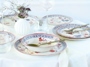 Gedeckter Tisch mit Porzellan-Tellern und Schalen im Blumenmuster in Blau und Rot, goldfarbenem Besteck, Glasvasen und Weingläsern, fotografiert aus leicht erhöhter schräger Perspektive (3/4-Ansicht).