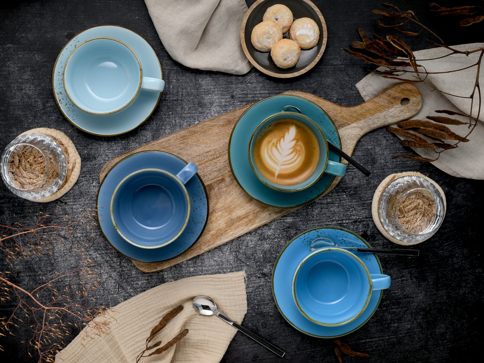 Draufsicht auf ein blaues Kaffeetassen- und Untertassen-Set auf dunklem Tisch, mit Latte Art in einer Tasse auf Holzbrett, Löffeln, Wassergläsern und kleinen Keksen.