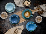 Draufsicht auf ein blaues Kaffeetassen- und Untertassen-Set auf dunklem Tisch, mit Latte Art in einer Tasse auf Holzbrett, Löffeln, Wassergläsern und kleinen Keksen.