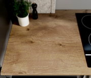 Close-up schuin bovenaanzicht van een houten keukenwerkblad met natuurlijke nerf en knoesten, naast een zwarte kookplaat; op de achtergrond een kruidenpot en pepermolen.