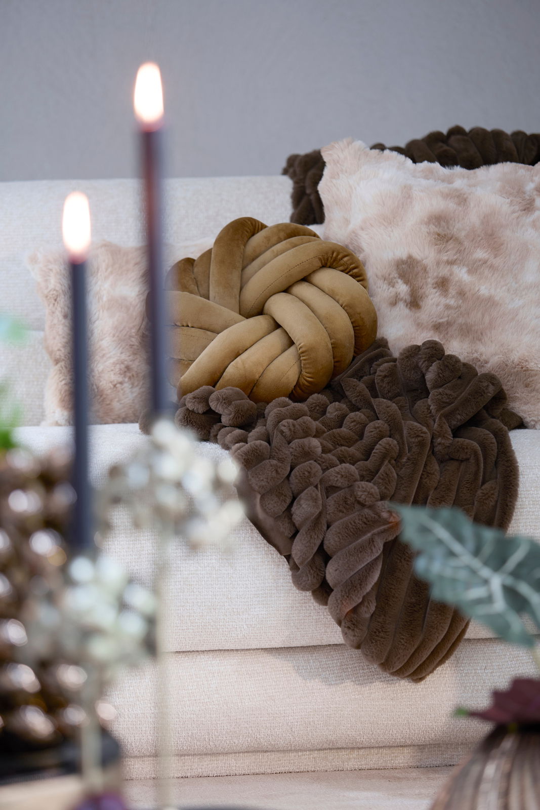 Ein gemütliches Wohnzimmer mit einem beigen Sofa, dekoriert mit einem geflochtenen, senfgelben Kissen und einem flauschigen, braunen Kissen. Im Vordergrund sind unscharfe Kerzen zu sehen. Perspektive: Frontalansicht.