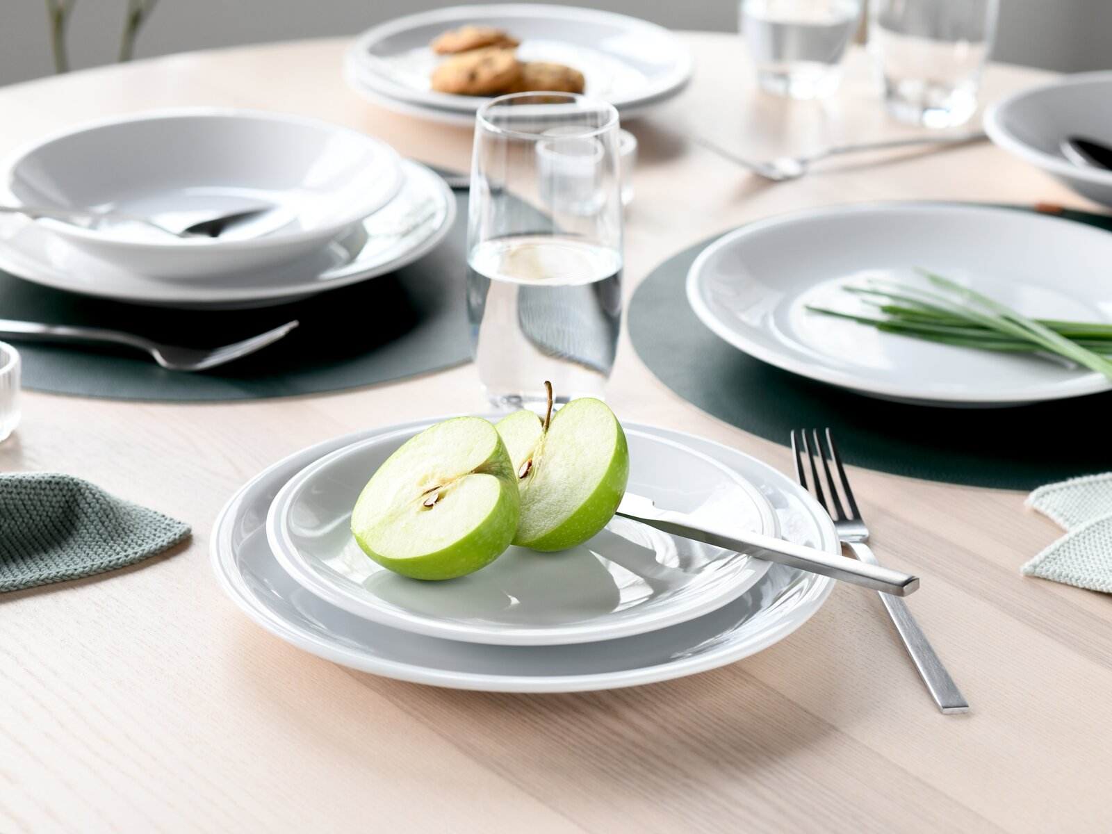 Modern gedeckter Esstisch mit weißem Geschirr, Besteck und Wassergläsern; vorn ein halbierter grüner Apfel auf einem kleinen Teller – Perspektive: leicht erhöhte, schräge Nahaufnahme von vorne.