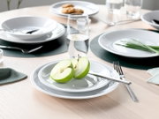 Modern gedeckter Esstisch mit weißem Geschirr, Besteck und Wassergläsern; vorn ein halbierter grüner Apfel auf einem kleinen Teller – Perspektive: leicht erhöhte, schräge Nahaufnahme von vorne.