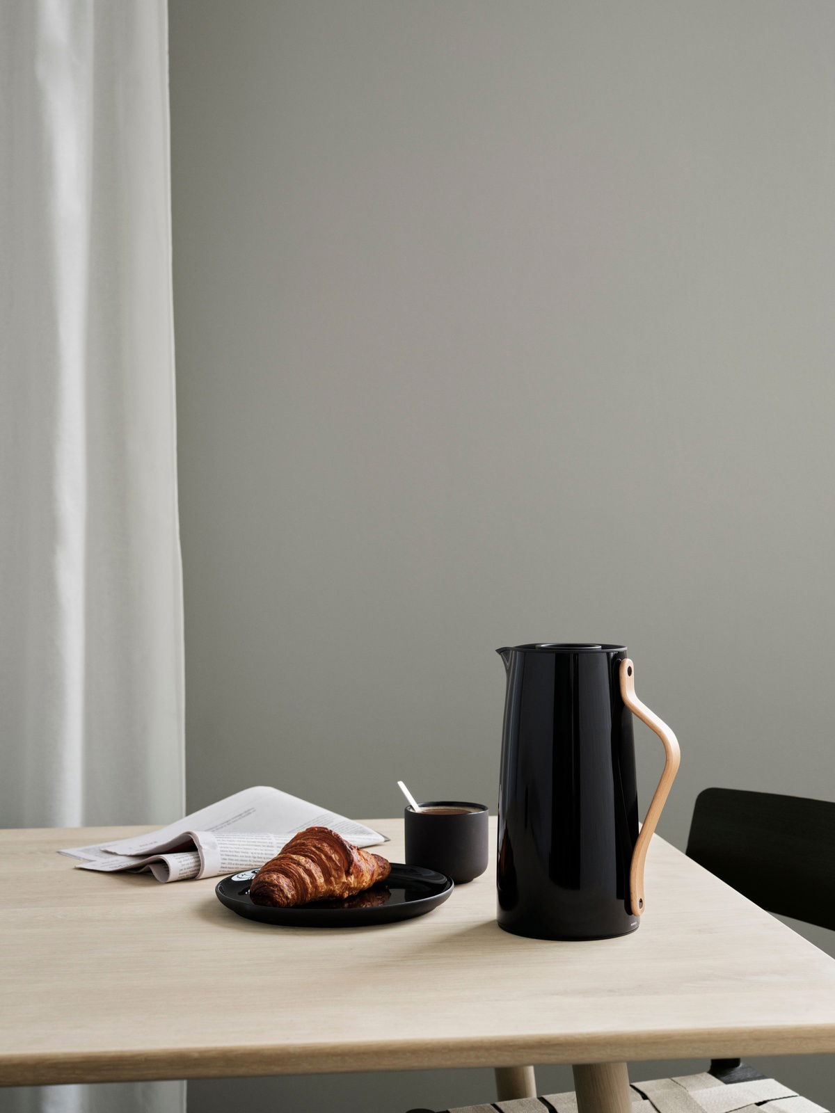 Schwarze Emma Isolierkanne von Stelton mit Holzgriff auf einem Tisch, neben einer Tasse Kaffee und einem Croissant, seitliche Perspektive.