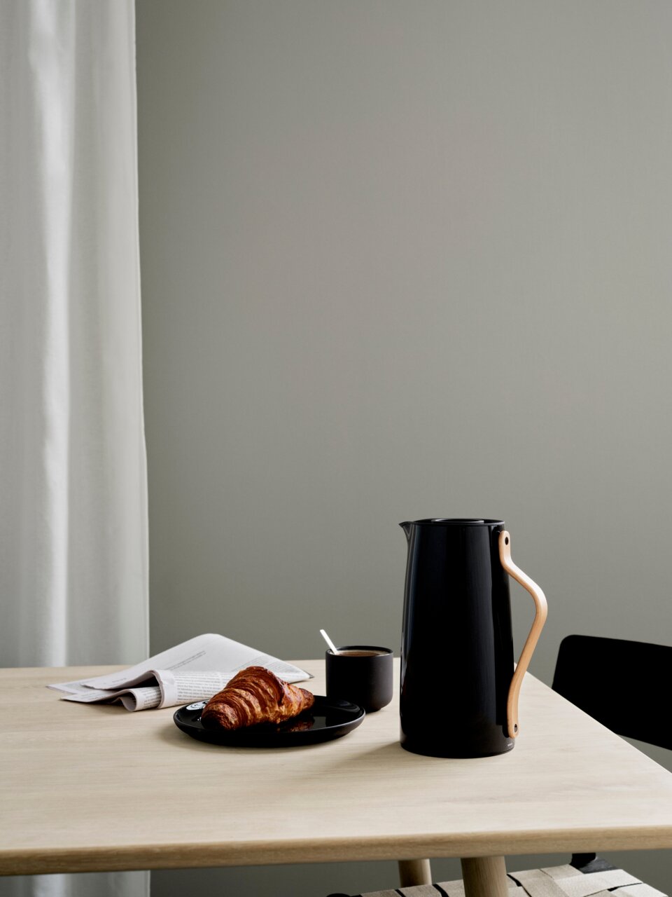 Zwarte Emma-thermoskan van Stelton met houten handvat op een tafel, naast een kopje koffie en een croissant, zijaanzicht.