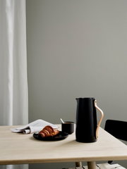 Zwarte Emma-thermoskan van Stelton met houten handvat op een tafel, naast een kopje koffie en een croissant, zijaanzicht.