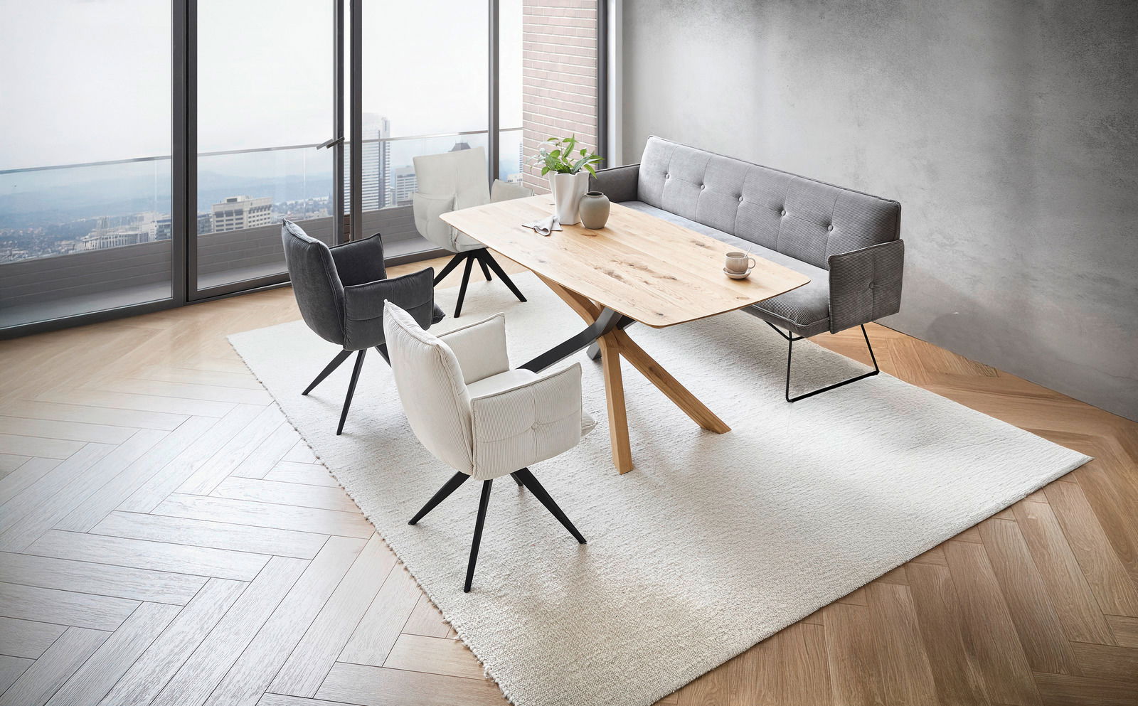Moderne Esszimmermöbel in einem hellen Raum mit Blick auf die Stadt, bestehend aus einem Holztisch, grauen und weißen Armlehnstühlen sowie einer grauen Bank. Perspektive von schräg oben.