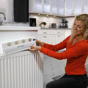 Frau installiert Invictus Therma Boost Heizgerät an einem Heizkörper in einer modernen Küche, seitliche Perspektive