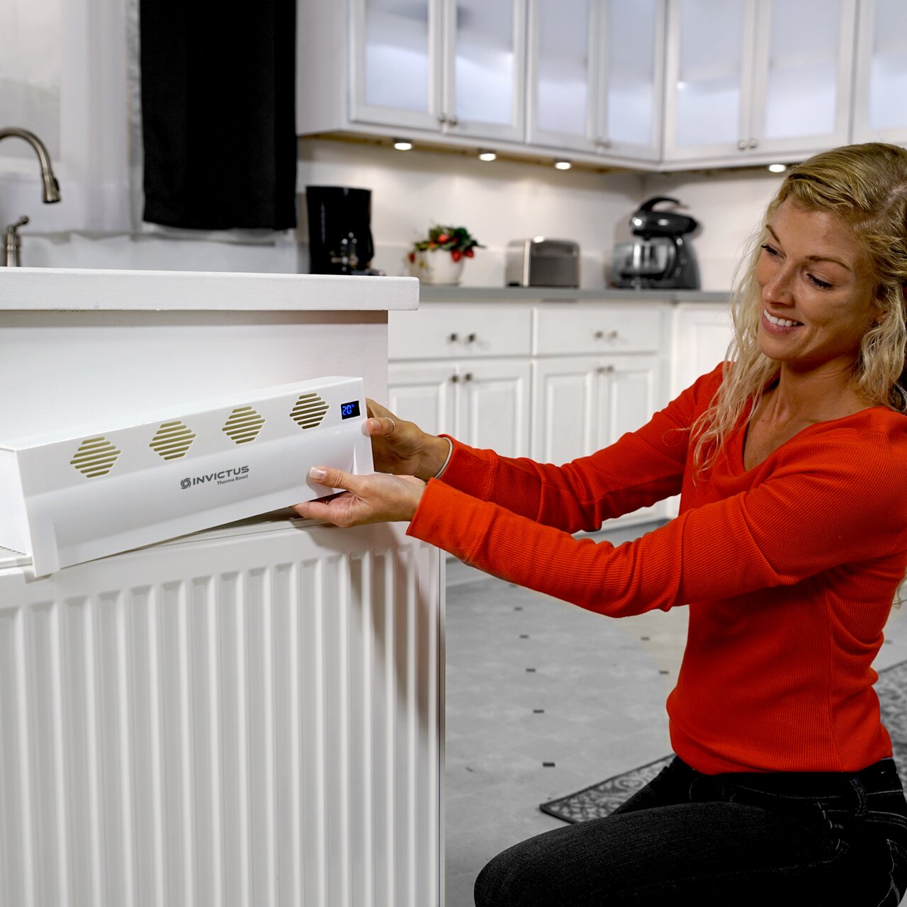 Een vrouw installeert een Invictus Therma Boost-verwarmingselement op een radiator in een moderne keuken, zijaanzicht