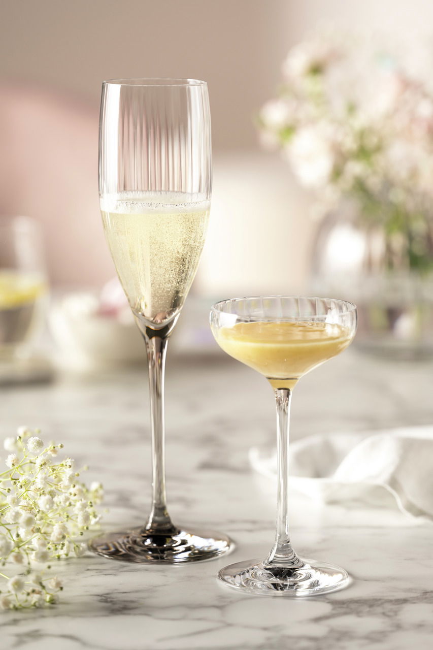 Zwei elegante Sektgläser auf einem Marmortisch, eines gefüllt mit Sekt, im Hintergrund unscharfe Blumen und weiche Beleuchtung, seitliche Perspektive.