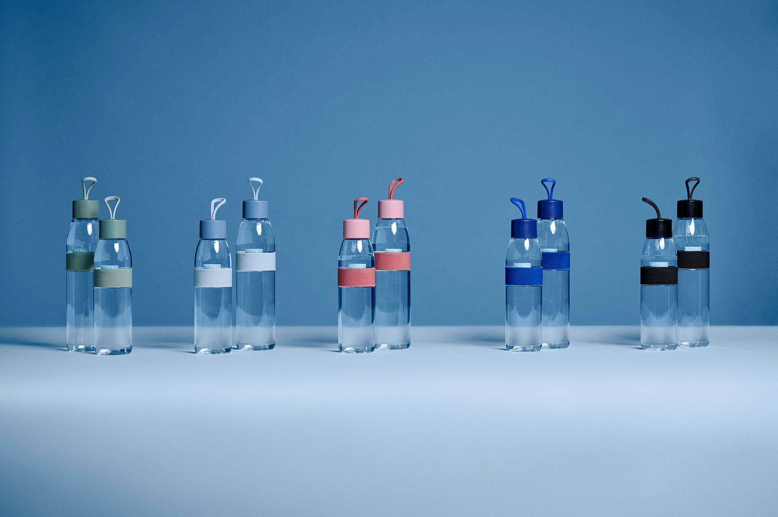 Mepal Trinkflasche Nordic blue ELLIPSE Mehrere transparente Trinkflaschen mit bunten Deckeln und Bändern, frontal auf einem blauen Hintergrund präsentiert.
