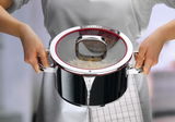 Person gießt Wasser aus einem glänzenden Kochtopf mit Glasdeckel ab, Vorderansicht.