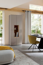 Moderne hoge opbergkast in matgrijs met open nis in houtlook en glazen plank, gefotografeerd in een lichte woonkamer vanuit een schuin vooraanzicht.