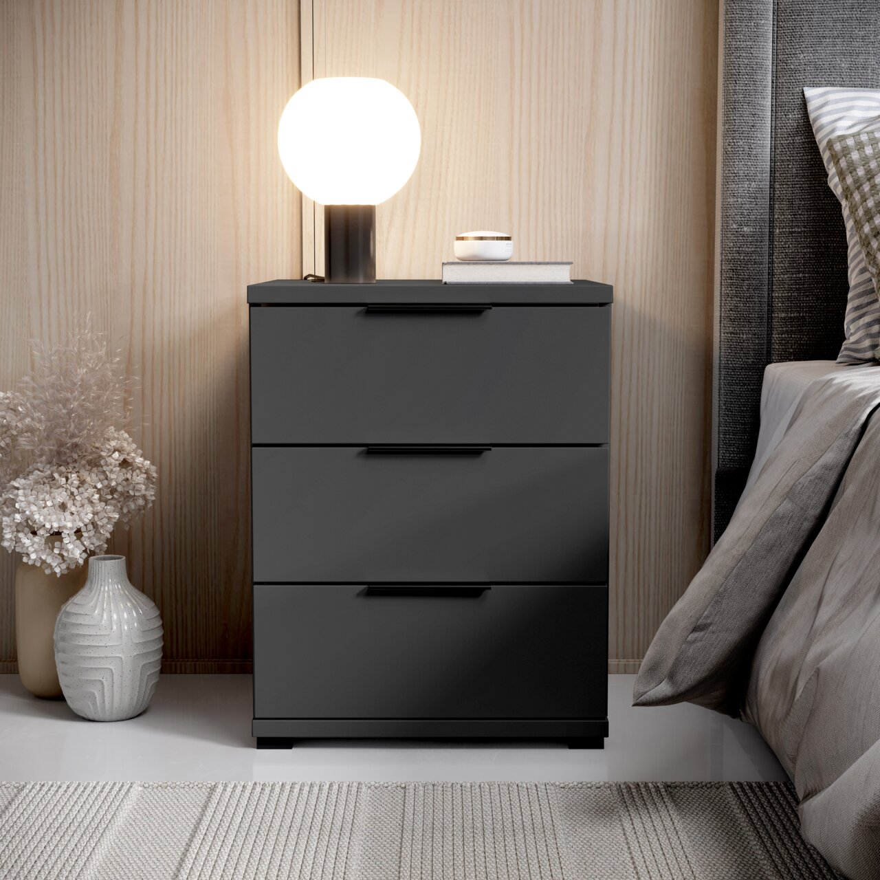 Z2 Nachtkommode SMILLA 2 Moderne Nachtkommode mit drei Schubladen in dunklem Design, seitliche Perspektive, dekoriert mit Lampe und Vasen.