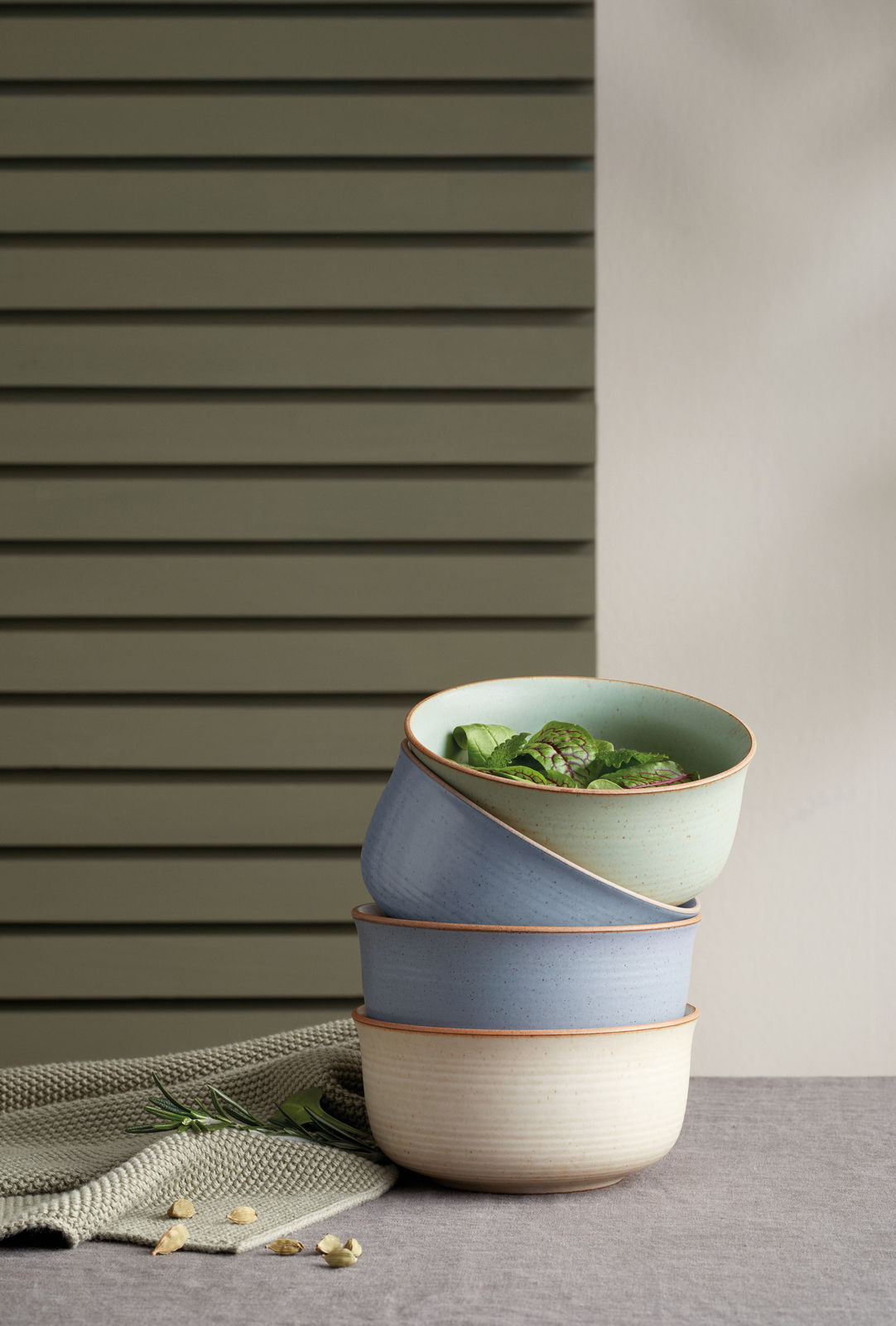 Stapel von drei Müsli-Schalen in verschiedenen Farben, von der Seite fotografiert, auf einem Tisch mit einem grünen Tuch und Kräutern.