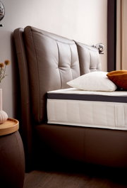 Nahaufnahme eines Boxspringbetts mit braunem, gepolstertem Kopfteil und weißer Matratze, seitliche Perspektive.