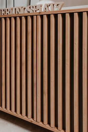 Nahaufnahme einer Kommode mit vertikalen Holzlatten und dekorativem Schriftzug auf der Oberseite, seitliche Perspektive