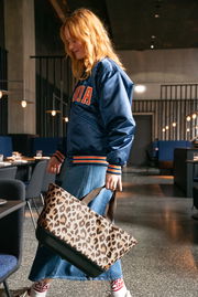 Frau in blauer Jacke und Jeansrock trägt einen Einkaufskorb mit Leopardenmuster in einem modernen Restaurant, seitliche Perspektive.