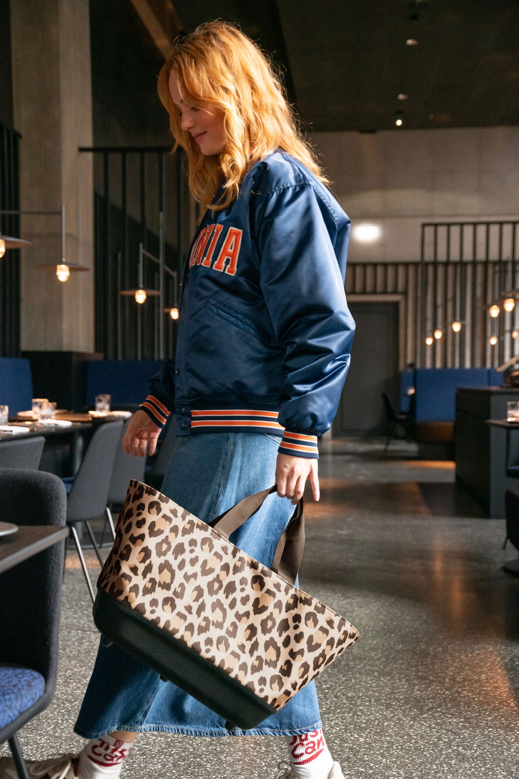 Frau in blauer Jacke und Jeansrock trägt einen Einkaufskorb mit Leopardenmuster in einem modernen Restaurant, seitliche Perspektive.
