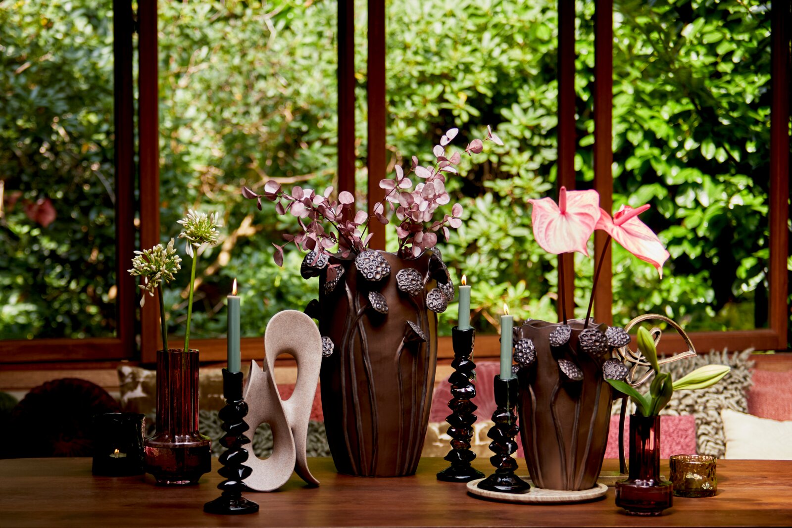 Vooraanzicht van een sfeervolle interieursetting met sculpturale bruine vazen, roze bloemen en decoratieve kandelaars op een houten tafel.