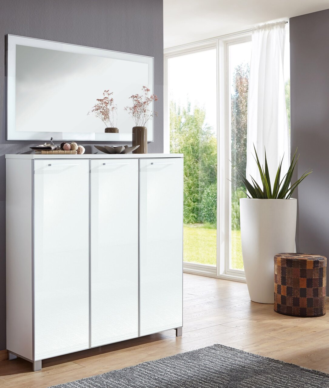 VOSS XL-Schuhschrank SANTINA Moderner XL-Schuhschrank in Weiß mit drei Türen, dekoriert mit Vasen, vor einem großen Fenster in einem hellen Raum, seitliche Perspektive.
