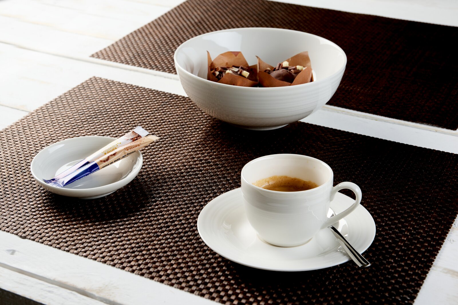 Weißes Porzellan-Kaffeegeschirr mit Tasse, Untertasse, Schälchen und großer Dessertschale auf braunen Tischsets, aus schräger Perspektive von vorn oben präsentiert.