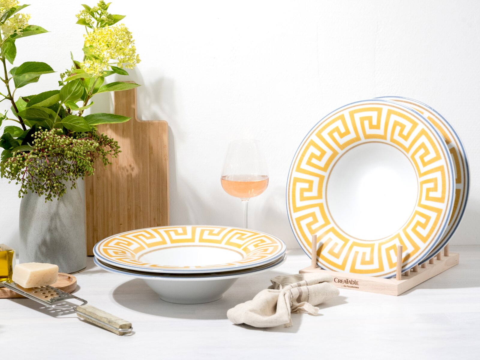 Creatable Pastateller 4-tlg GOURMET Weißes Geschirrset mit gelb-goldenem Mäander-Muster und blauer Kante, Teller im Ständer und tiefer Teller im Vordergrund, fotografiert in leichter Seitenansicht auf weißem Tisch.