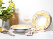 Weißes Geschirrset mit gelb-goldenem Mäander-Muster und blauer Kante, Teller im Ständer und tiefer Teller im Vordergrund, fotografiert in leichter Seitenansicht auf weißem Tisch.