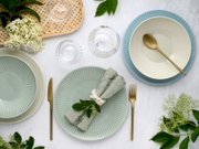 Pastellfarbenes Geschirr-Set mit geriffelten Tellern und Schalen (Mint, Creme, Blau), goldfarbenem Besteck, Leinenserviette und Glas auf hellem Tisch, Draufsicht von oben