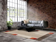 Moderner Wohnzimmerteppich Castle Flora in Beige mit roten und braunen Akzenten, ca. 200x250cm, vor einer Ledercouch und einem Couchtisch in einem industriellen Loft mit Backsteinwand und großen Fenstern.