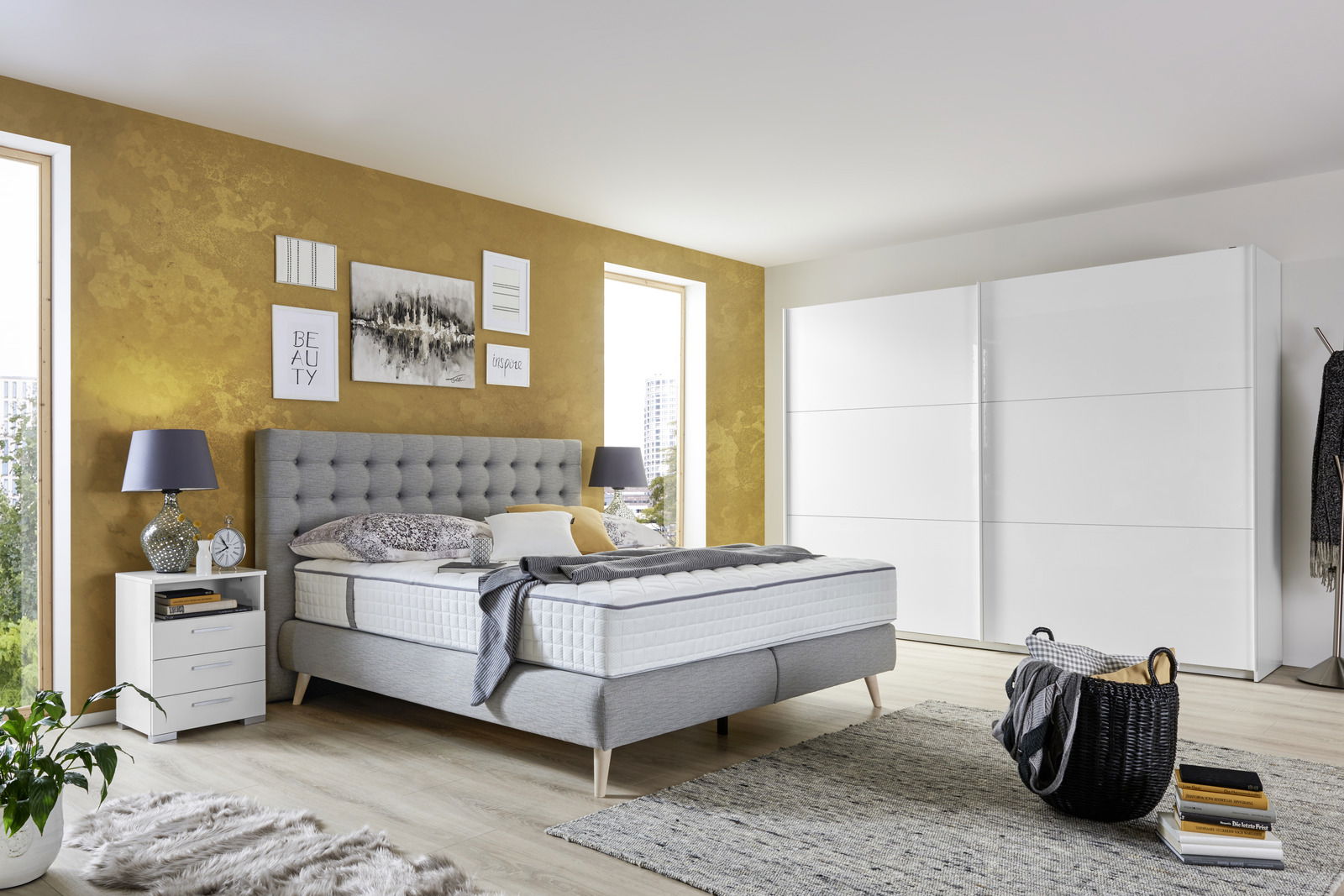 Modernes Boxspringbett in einem stilvollen Schlafzimmer mit gelber Akzentwand, grauem Kopfteil und weißem Nachttisch, seitliche Perspektive.