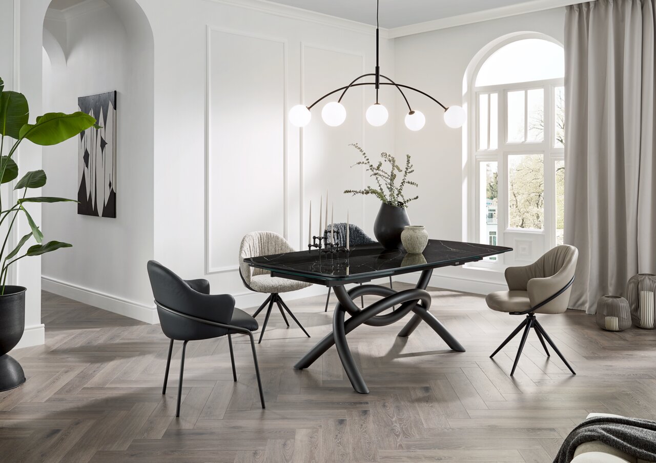 Belluti Armlehnstuhl MATTEO Moderner Essbereich mit einem schwarzen Esstisch mit geschwungenem Untergestell und verschiedenen Armlehnstühlen in einem hellen Raum mit großen Fenstern, aus der Perspektive von schräg vorne.