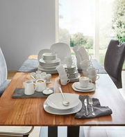 Ein 62-teiliges Kombiservice-Set aus weißem Porzellan, präsentiert auf einem Holztisch in einem modernen Esszimmer mit Blick auf den Garten. Die Perspektive zeigt das Set von schräg oben, mit Tellern, Tassen, Schalen und Besteck, die elegant arrangiert si