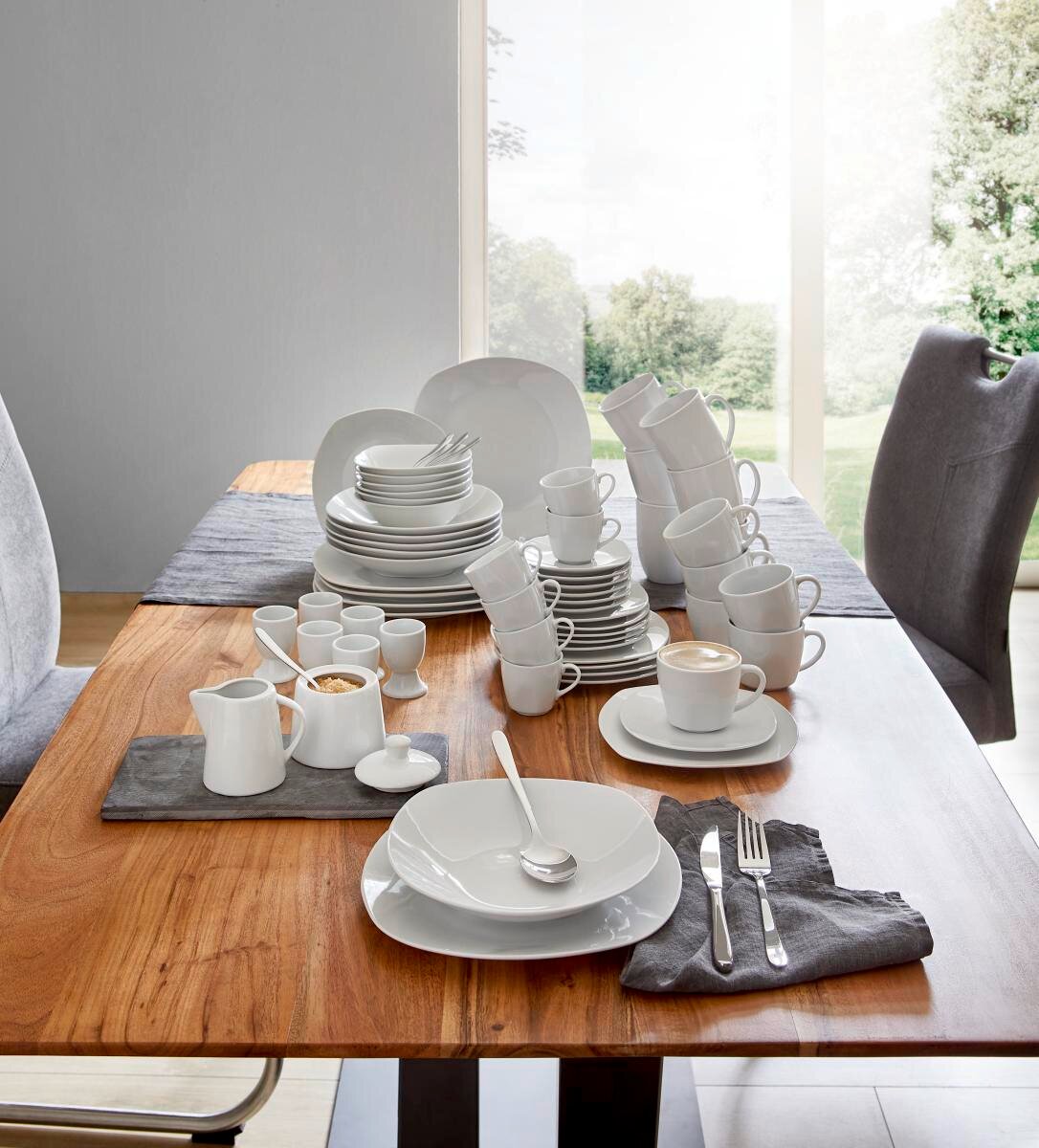Een 62-delige servieset van wit porselein, gepresenteerd op een houten tafel in een moderne eetkamer met uitzicht op de tuin. Het perspectief toont de set vanuit een schuine hoek van bovenaf, met borden, kopjes, schalen en bestek die elegant zijn gerangsc