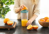 Frau presst eine Orange mit einem grünen Küchenhelfer auf einem Tisch aus, seitliche Perspektive.