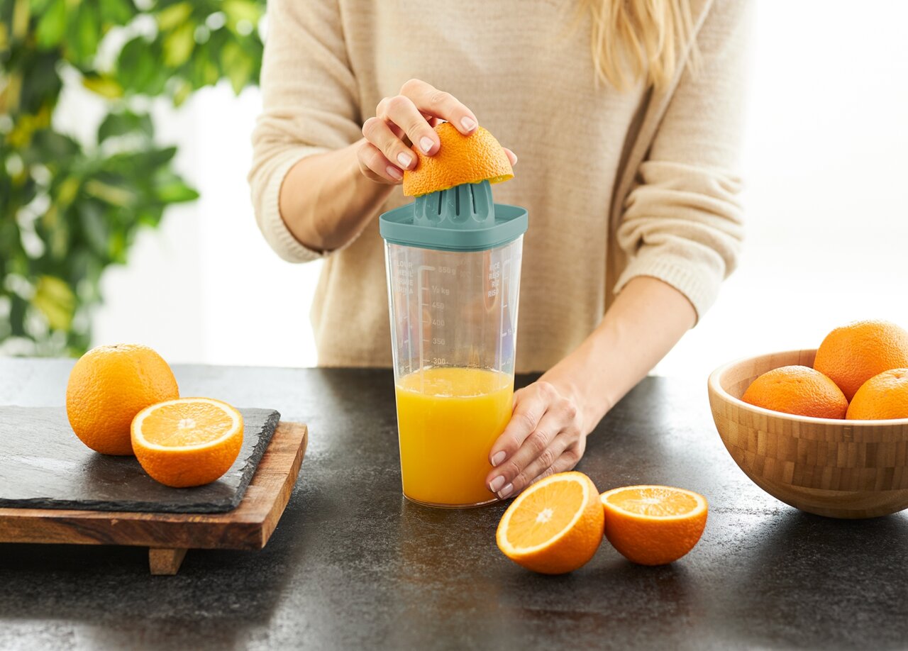 rotho Shake LOFT Frau presst eine Orange mit einem grünen Küchenhelfer auf einem Tisch aus, seitliche Perspektive.