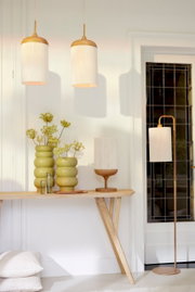 Een lichte woonruimte met twee cilindervormige hanglampen van lichte stof en hout, een bijpassende tafellamp op een houten voet en een staande lamp naast het raam; vooraanzicht van de lampen